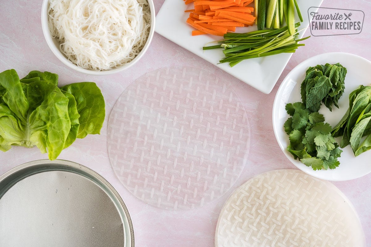 Rice paper wrappers, lettuce, matchstick veggies, fresh herbs, and rice noodles all laid out on the counter for fresh spring rolls.