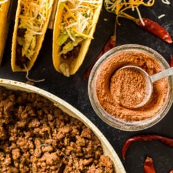 Taco seasoning in a glass jar next to tacos and taco meat in a pan