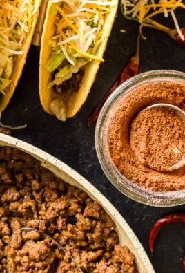 Taco seasoning in a glass jar next to tacos and taco meat in a pan