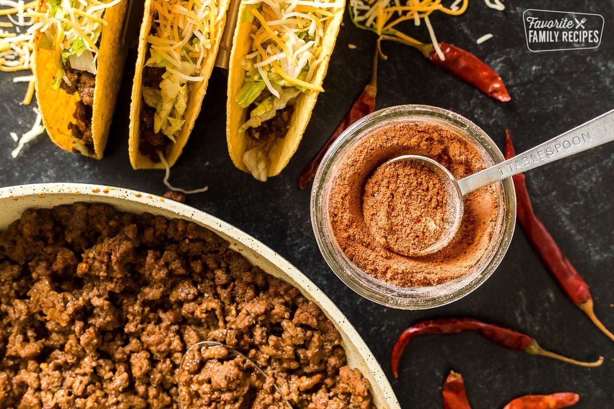 Taco seasoning in a glass jar next to tacos and taco meat in a pan.