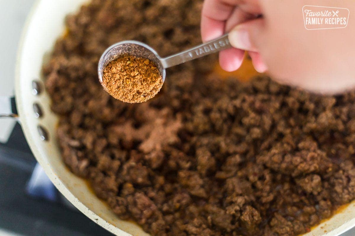 Homemade taco seasoning being spooned into a pan of taco meat