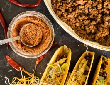 Three tacos, ground beef taco meat, and taco seasoning in a jar on a black table top.
