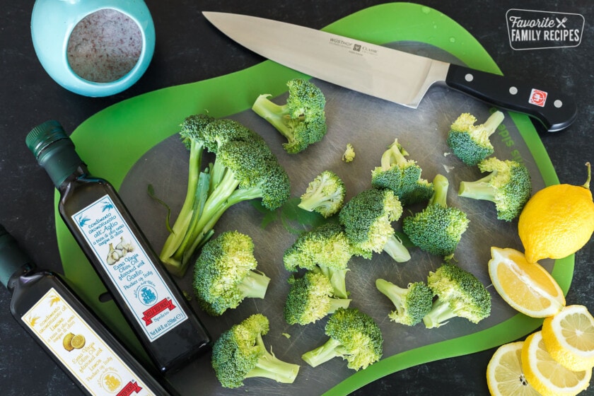 Cut up pieces of broccoli on a cutting board next to a knife, lemons, salt, and olive oil