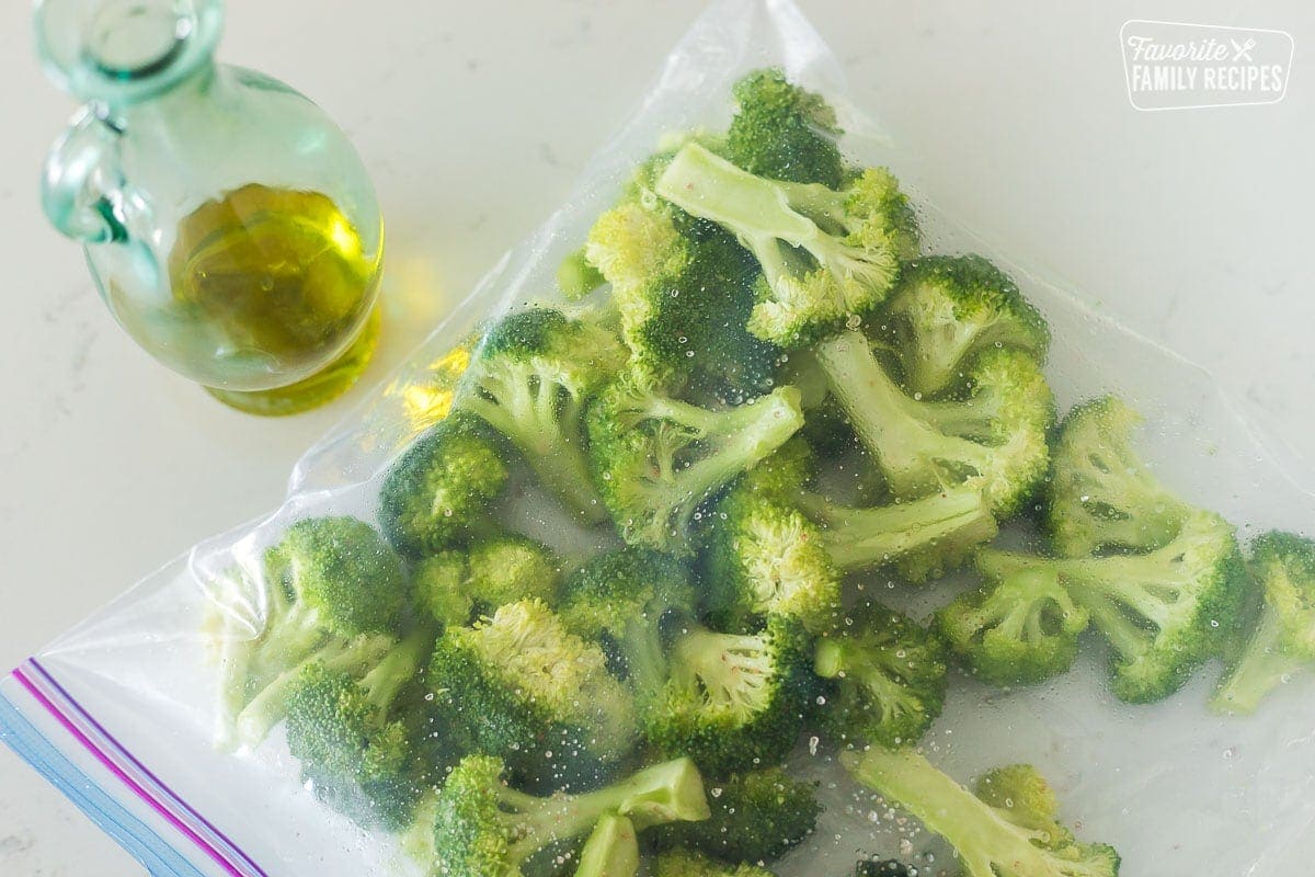 Raw broccoli cut into florets in a plastic zip-top bag next to a jar of olive oil