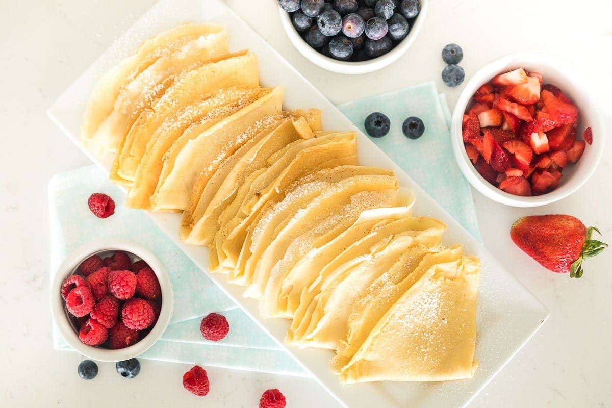 Plain homemade crepes on a white serving platter next to bowls of berries.