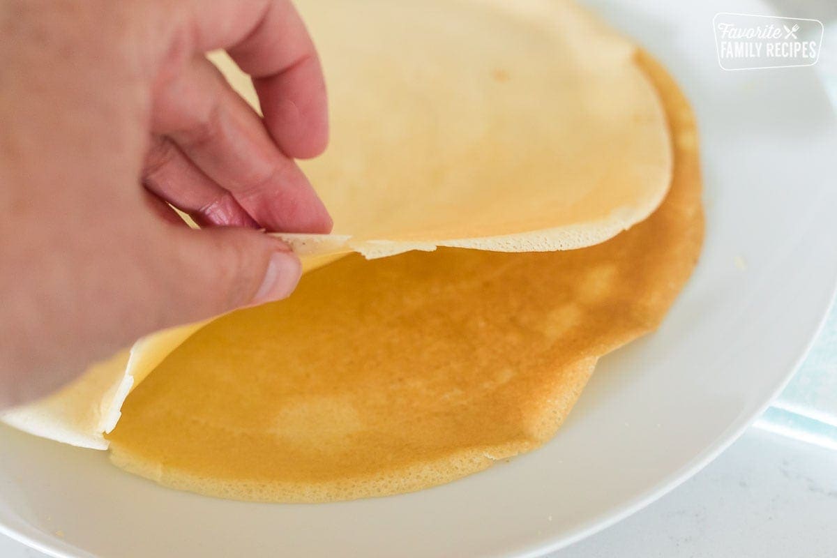 A hand lifting a crepe from s plate of crepes to show thin texture