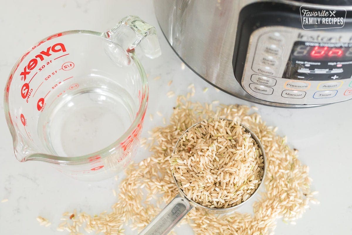 A liquid measuring cup with water and a cup of uncooked brown rice next to an Instant Pot
