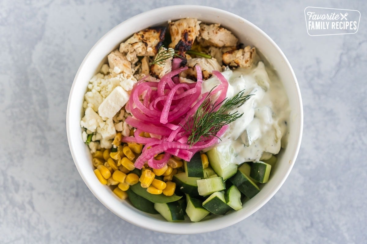 A Mediterranean bowl with saffron rice, spinach, chicken, corn, cucumbers, feta cheese, tzatziki sauce, pickled red onions, and fresh dill