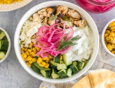 A Mediterranean bowl with saffron rice, spinach, chicken, corn, cucumbers, feta cheese, tzatziki sauce, pickled red onions, and fresh dill