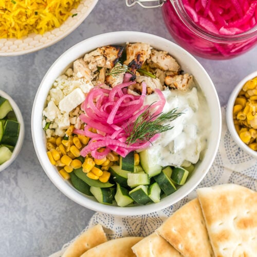 A Mediterranean bowl with saffron rice, spinach, chicken, corn, cucumbers, feta cheese, tzatziki sauce, pickled red onions, and fresh dill