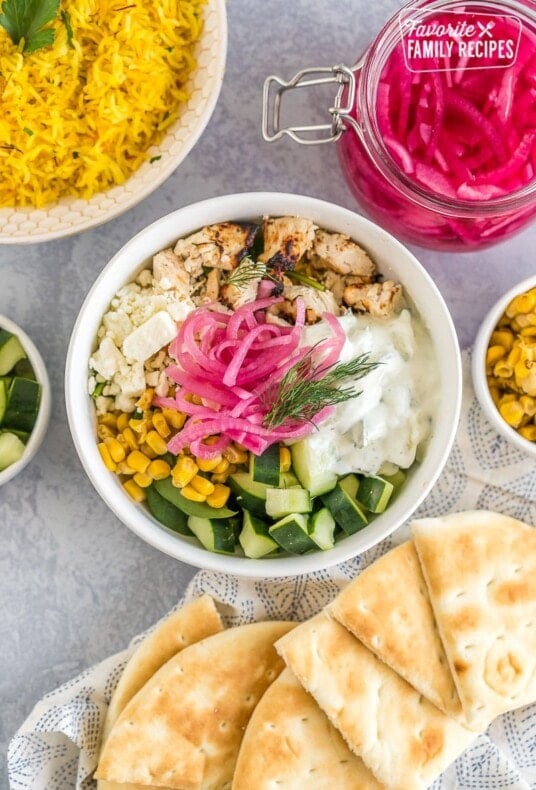 A Mediterranean bowl with saffron rice, spinach, chicken, corn, cucumbers, feta cheese, tzatziki sauce, pickled red onions, and fresh dill