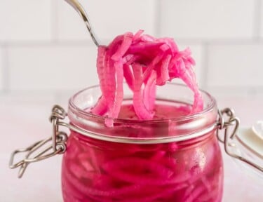 A hand using a fork to scoop some pickled red onions out of a jar