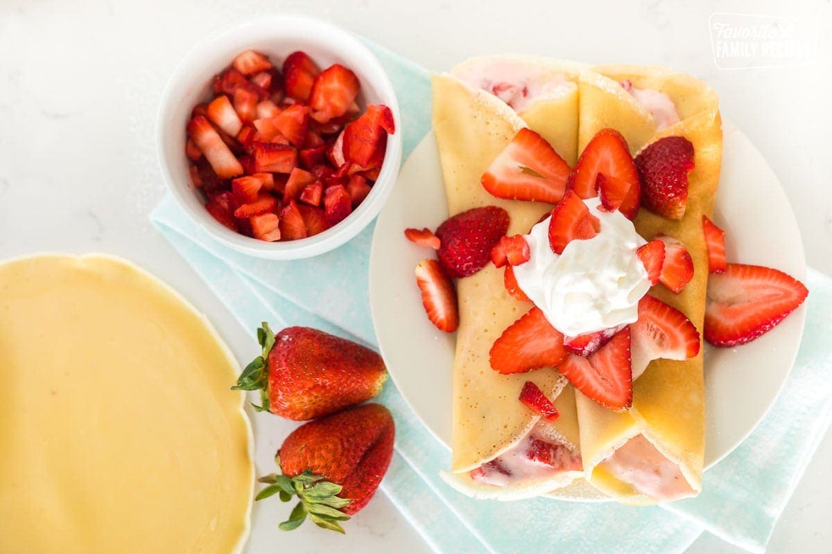 Two strawberry crepes next to a bowl of chopped strawberries and two whole strawberries