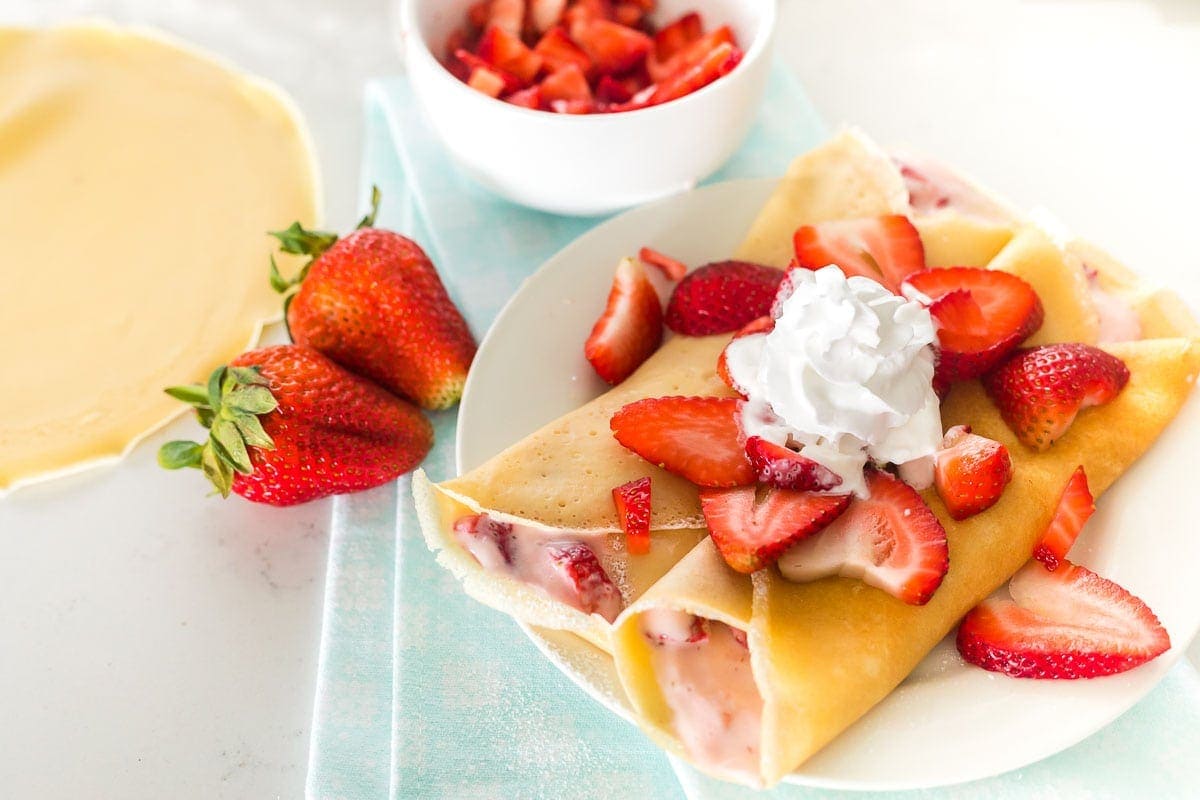 Two strawberry crepes on a plate topped with strawberries and whipped cream