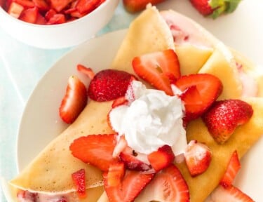 Two strawberry crepes on a plate topped with strawberries and whipped cream