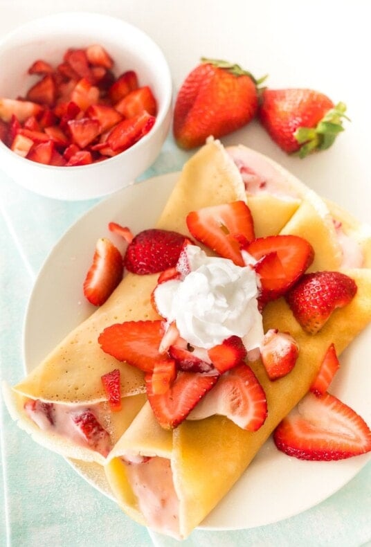 Two strawberry crepes on a plate topped with strawberries and whipped cream