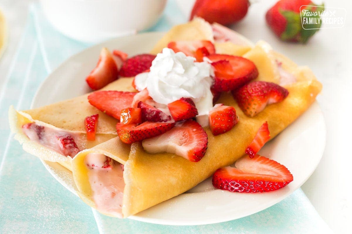 Two crepes on a white plate with strawberry filling and topped with whipped cream and strawberries