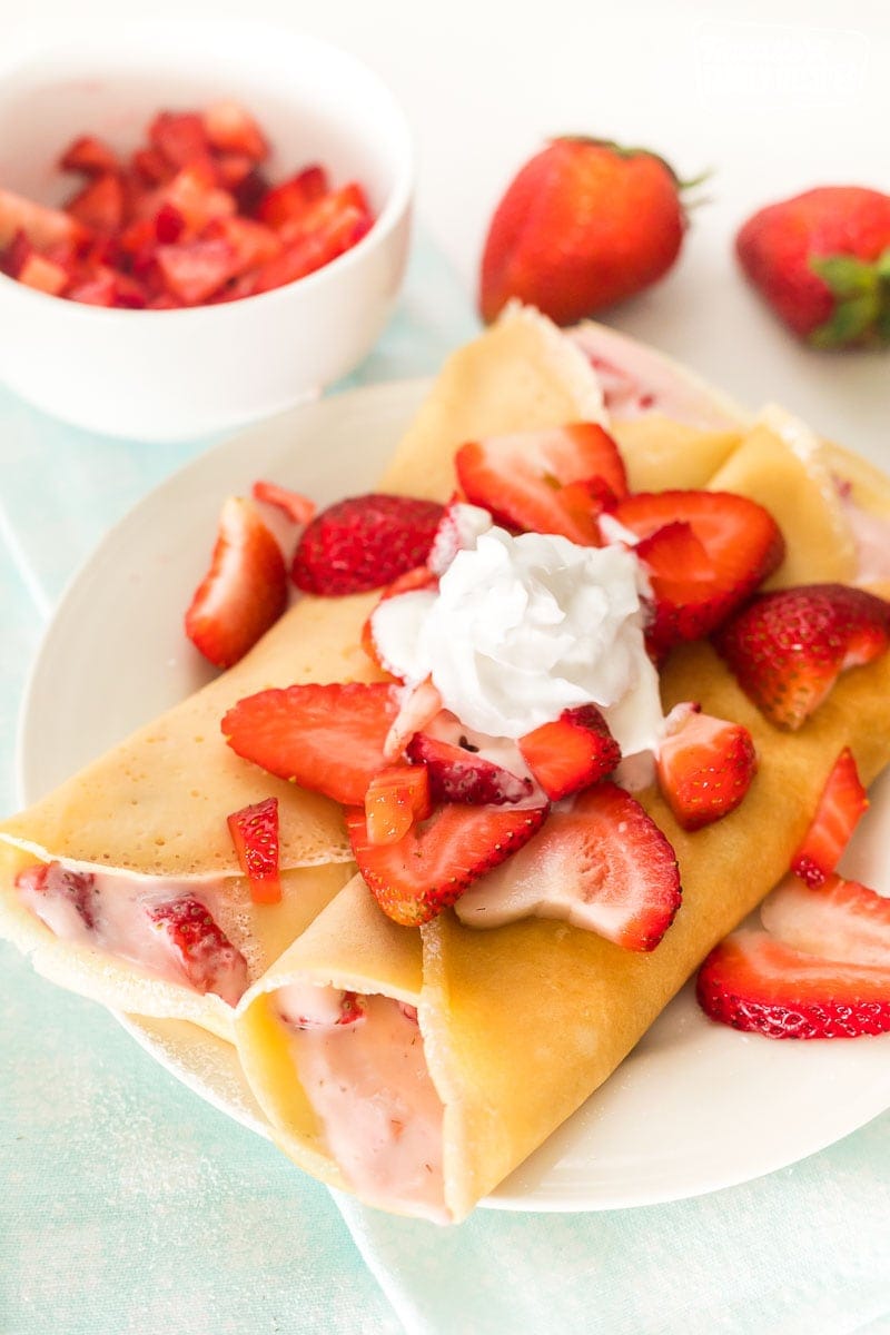 Two strawberry crepes on a plate topped with strawberries and whipped cream