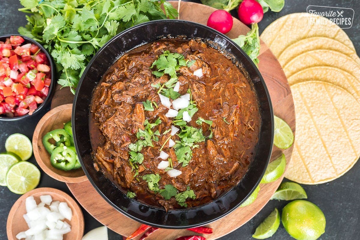 Top view of beef birria in a serving bowl next to jalapeños, limes, onion, radishes, and cilantro