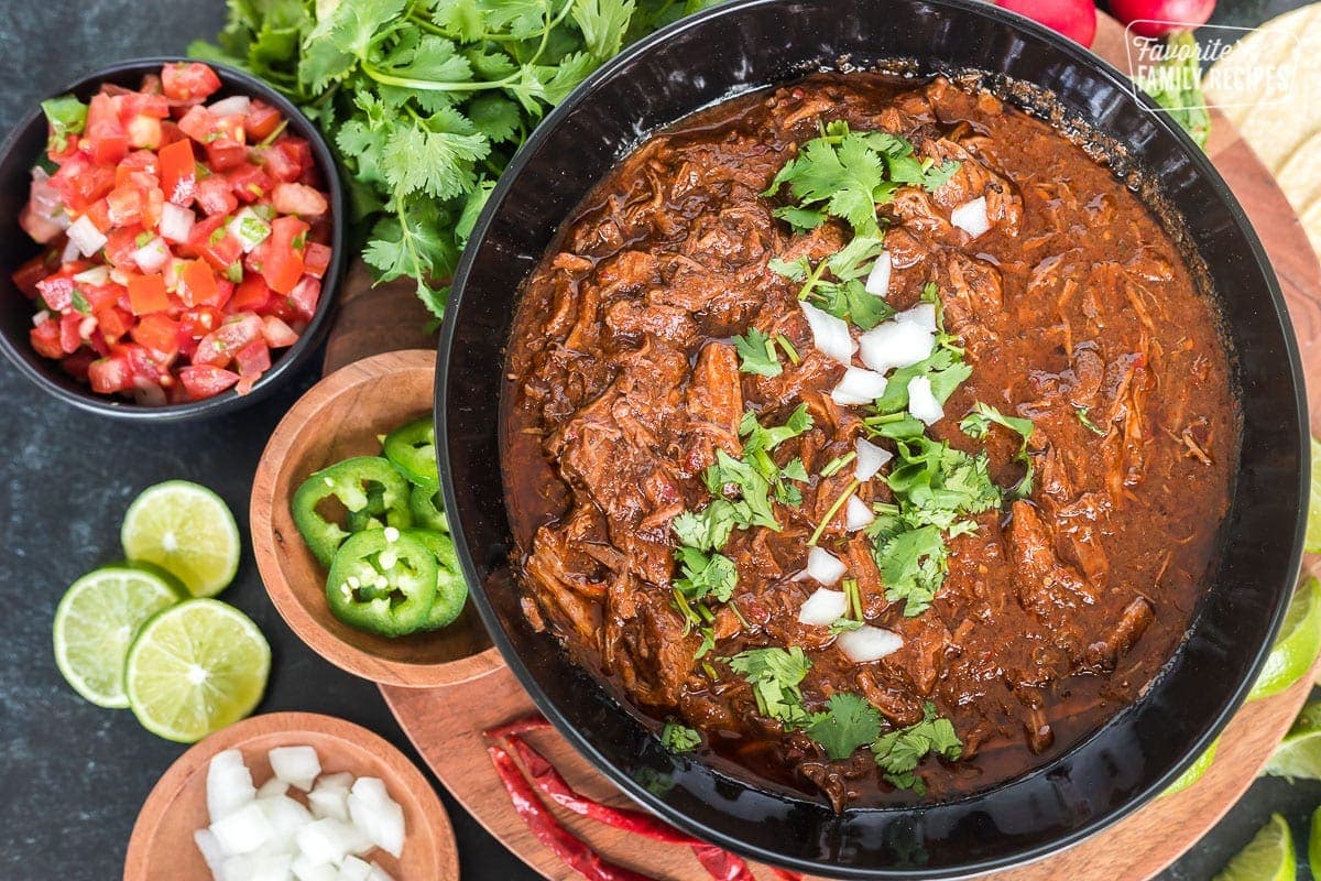 Braised and shredded beef birria in a black serving bowl on a wood platter, surrounded by tomatoes, sliced jalapenos, chopped onion, lime slices, and cilantro.