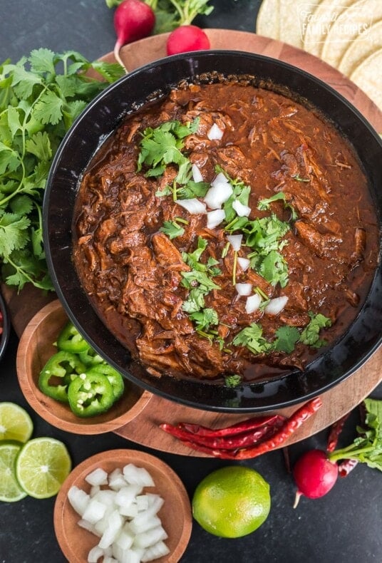 Top view of beef birria in a serving bowl next to jalapeños, limes, onion, radishes, and cilantro