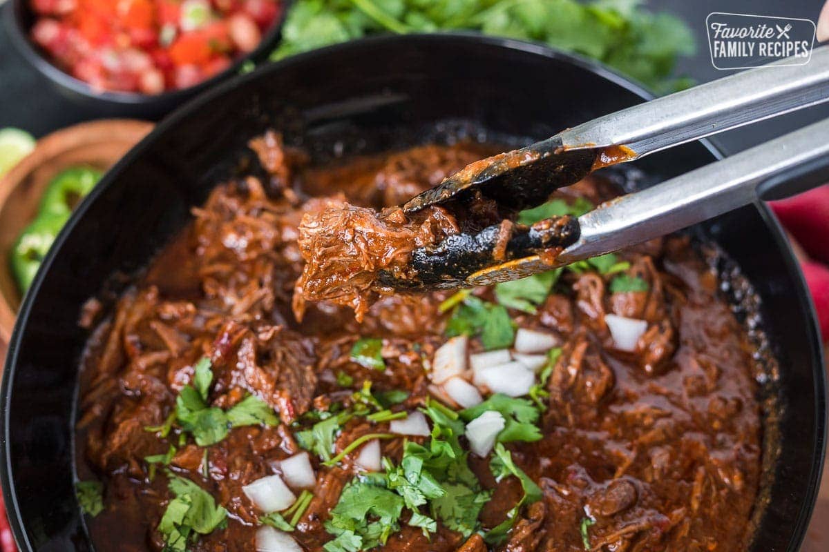 Tongs picking up a chunk of braised beef birria from a serving bowl