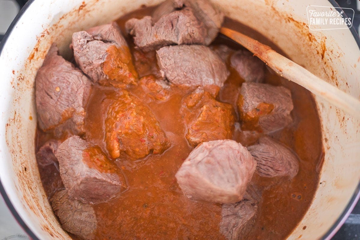 Pieces of chuck roast that have been browned and put in a Dutch oven. A red sauce is poured over the top