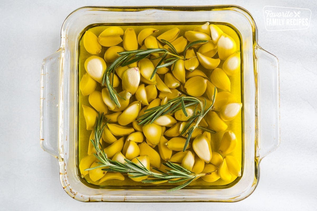 A baking dish full of whole peeled garlic cloves, olive oil, and rosemary sprigs