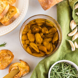 A jar of garlic confit surrounded by a baguette, rosemary, and garlic cloves