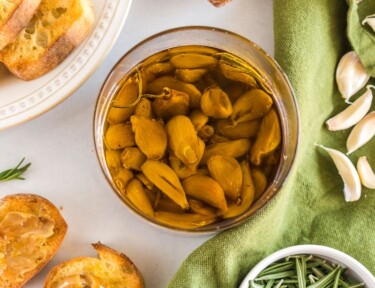 A jar of garlic confit surrounded by a baguette, rosemary, and garlic cloves