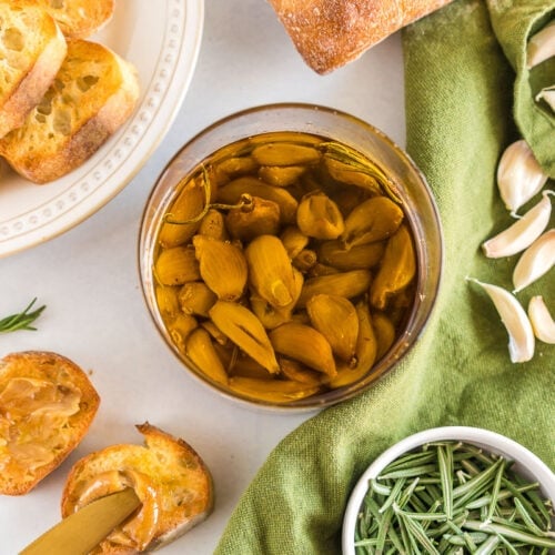 A jar of garlic confit surrounded by a baguette, rosemary, and garlic cloves