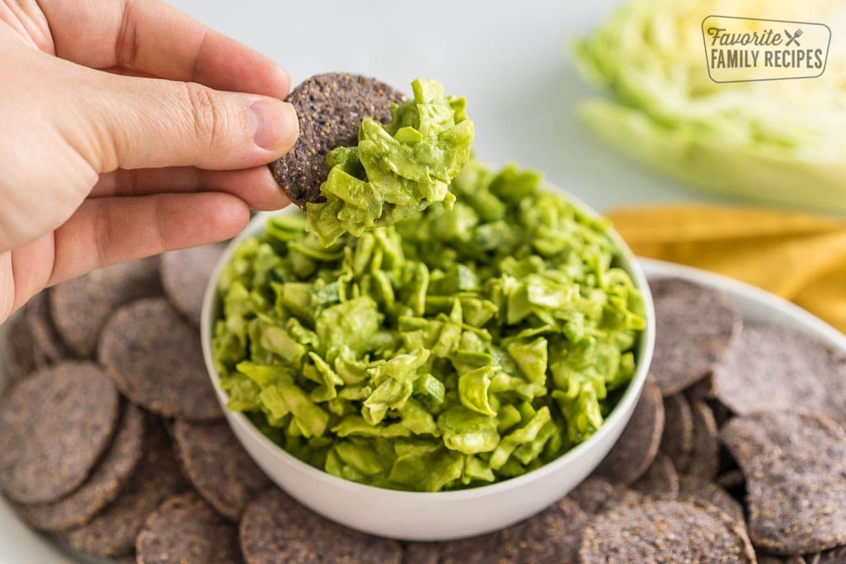A bowl of green goddess dip on a plate of blue corn chips with a hand holding a chip with dip on it.