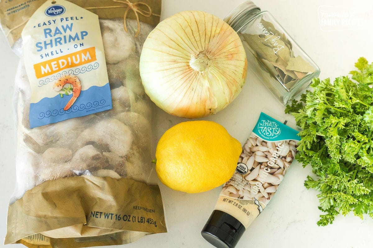 A bag of frozen, uncooked shrimp, an onion, a lemon, garlic, bay leaves, and parsley on a counter top.