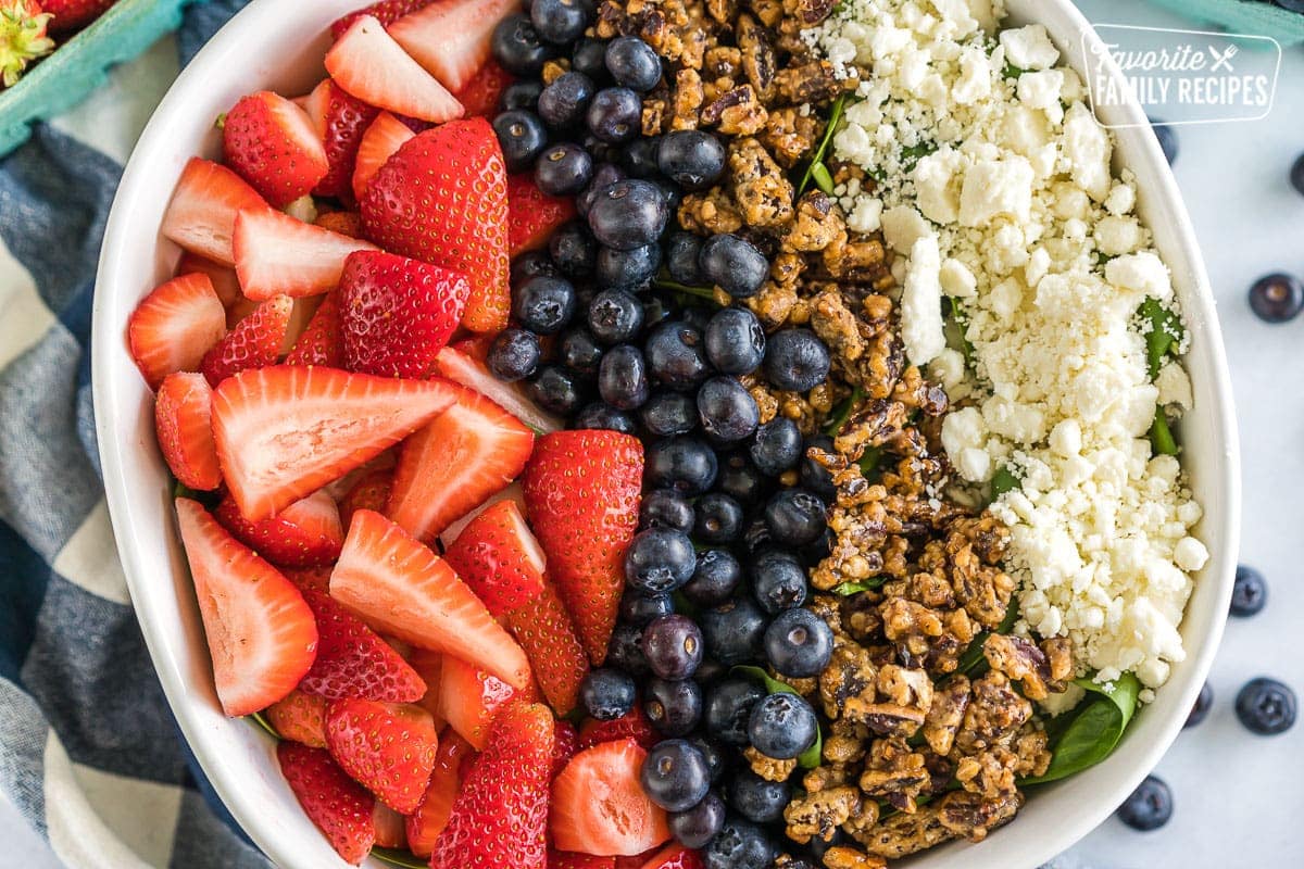 A large serving bowl filled with spinach and topped with chopped strawberries, blueberries, candied pecans, and feta cheese.