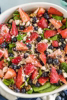 A large serving bowl filled with spinach and topped with chopped strawberries, blueberries, candied pecans, and feta cheese