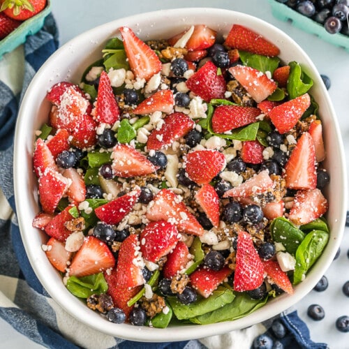 A large serving bowl filled with spinach and topped with chopped strawberries, blueberries, candied pecans, and feta cheese