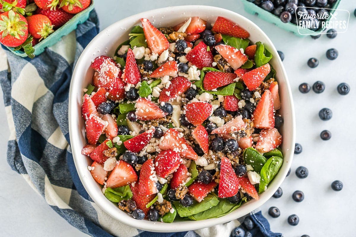 A large serving bowl filled with spinach and topped with chopped strawberries, blueberries, candied pecans, and feta cheese.