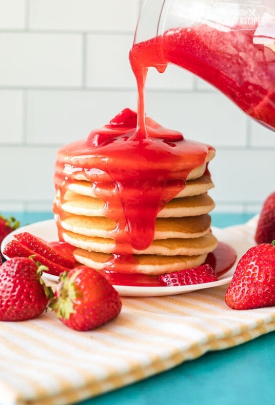 Strawberry syrup being poured over a large stack of pancakes