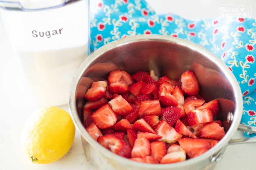 Fresh strawberries on a saucepan next to a container of sugar and a lemon