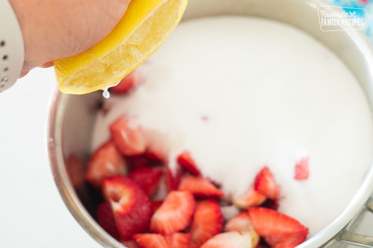 Strawberries and sugar in a saucepan with a lemon being squeezed over the top