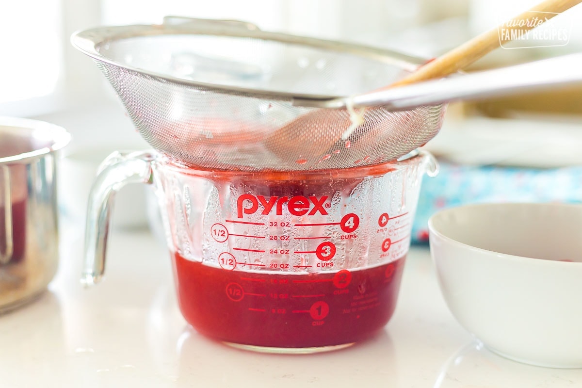 Cooked strawberries being pressed through a fine mesh strainer into a liquid measuring cup