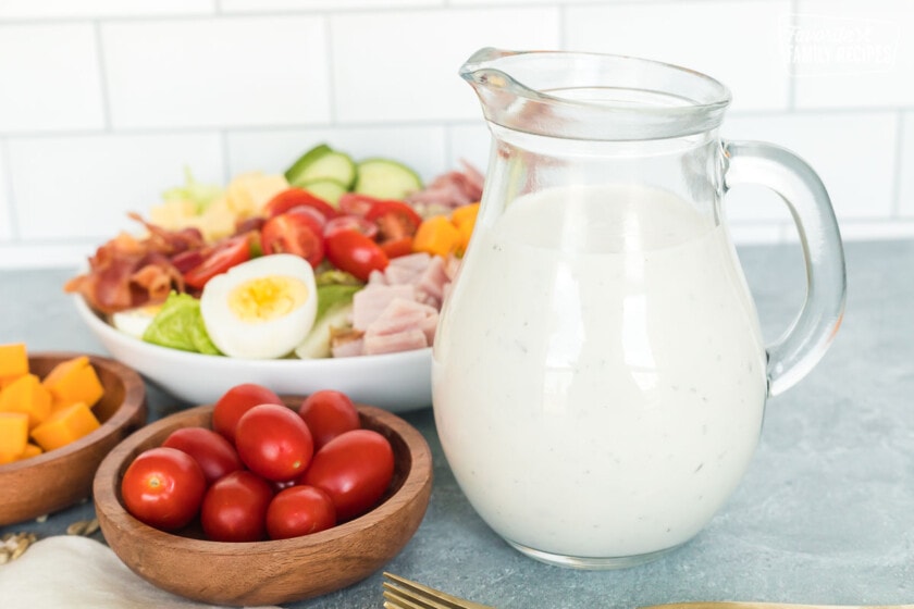 A clear salad dressing pitcher with homemade Ranch dressing next to a chef salad