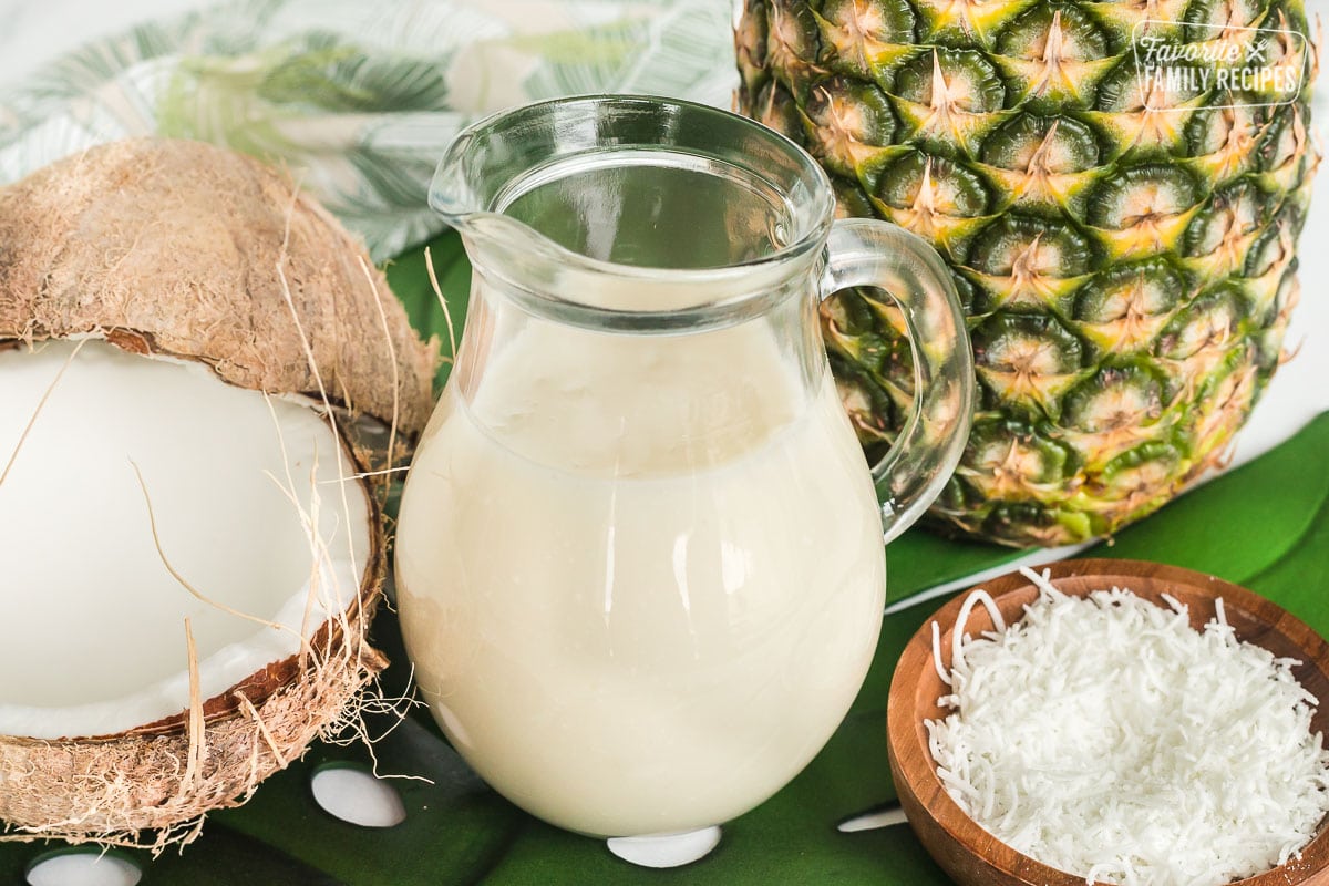 A clear, glass pitcher of coconut syrup next to a fresh coconut and a pineapple