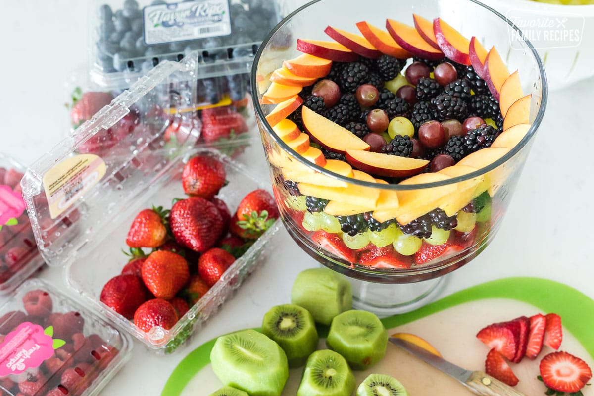 A partially made fruit trifle with cut up fruits next to it on a cutting board