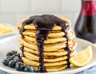 A stack of lemon ricotta pancakes on a plate covered in blueberry syrup with blueberries and a lemon wedge