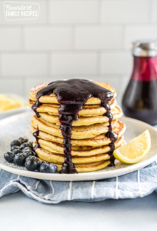 A stack of lemon ricotta pancakes on a plate covered in blueberry syrup with blueberries and a lemon wedge