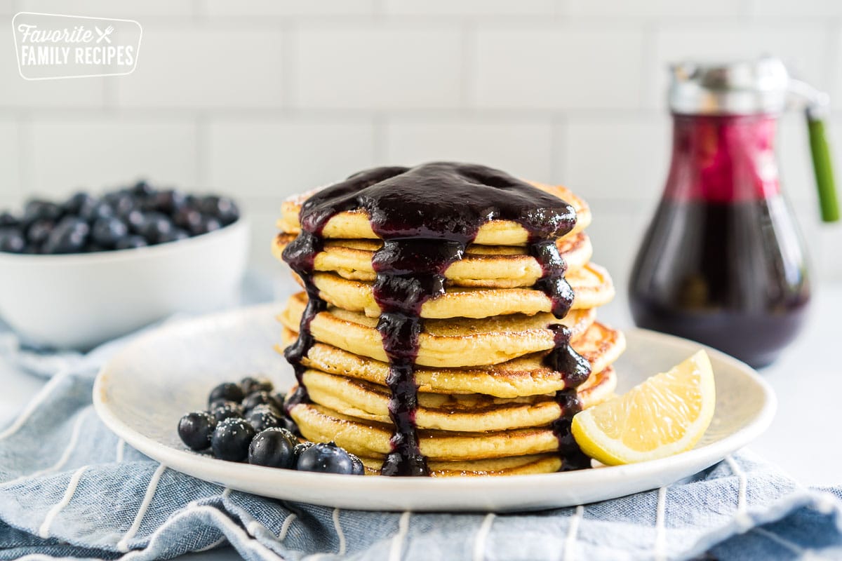 A stack of lemon ricotta pancakes on a plate covered in blueberry syrup with blueberries and a lemon wedge.