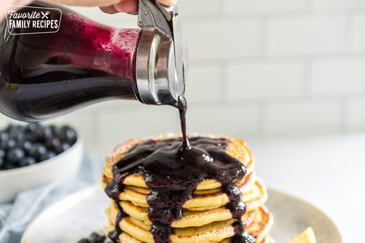 A syrup dispenser pouring blueberry syrup on a stack of pancakes.