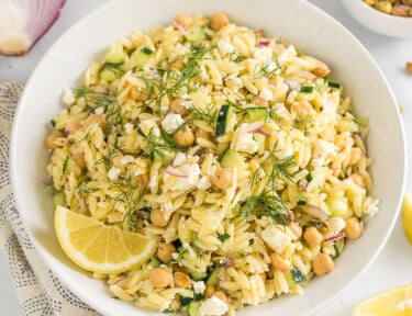 A large white bowl full of orzo salad garnished with fresh dill and a lemon wedge