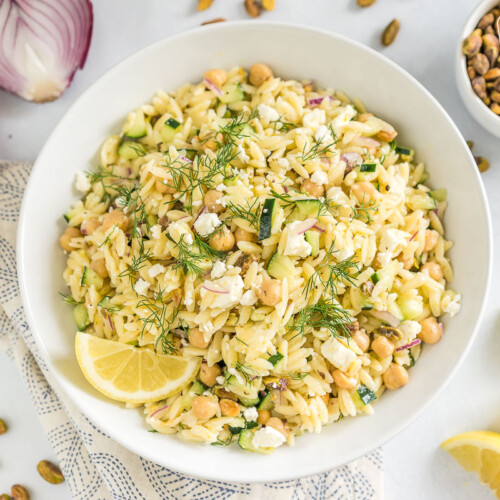 A large white bowl full of orzo salad garnished with fresh dill and a lemon wedge